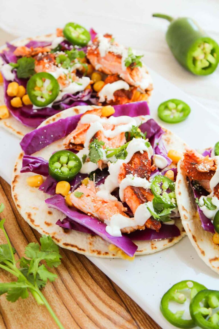 Three tacos with grilled salmon, purple cabbage, corn, jalapeño slices, and drizzled sauce on a white plate. A sliced jalapeño and cilantro garnish are nearby.