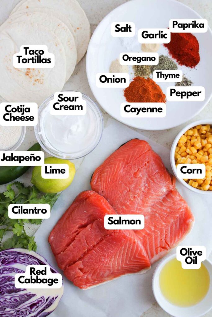 Ingredients for making salmon tacos are arranged, including salmon, taco tortillas, cilantro, limes, jalapeño, red cabbage, corn, olive oil, spices, sour cream, and cotija cheese.