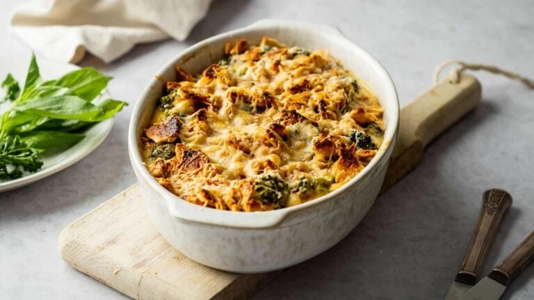 Baked casserole in a white dish on a wooden board, with melted cheese and vegetables, alongside a plate of fresh basil leaves.