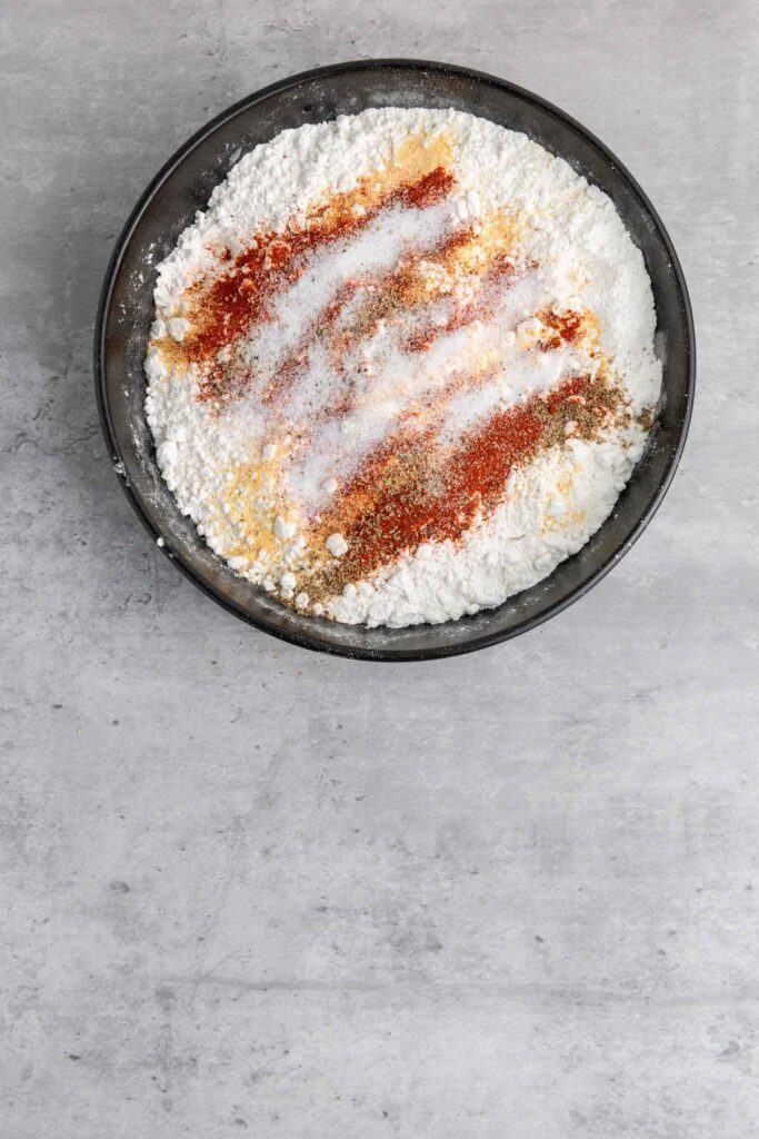A black bowl filled with white flour and an array of spices, including red, brown, and yellow powders, sits on a gray stone surface—perfect for crafting buttermilk fried chicken.