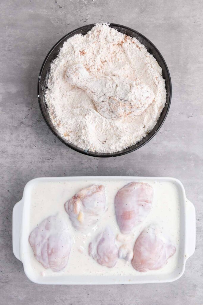 Chicken pieces marinating in buttermilk sit in a white dish with a milky liquid, next to a black bowl containing flour-coated chicken on a gray countertop, ready for that crispy fried chicken perfection.