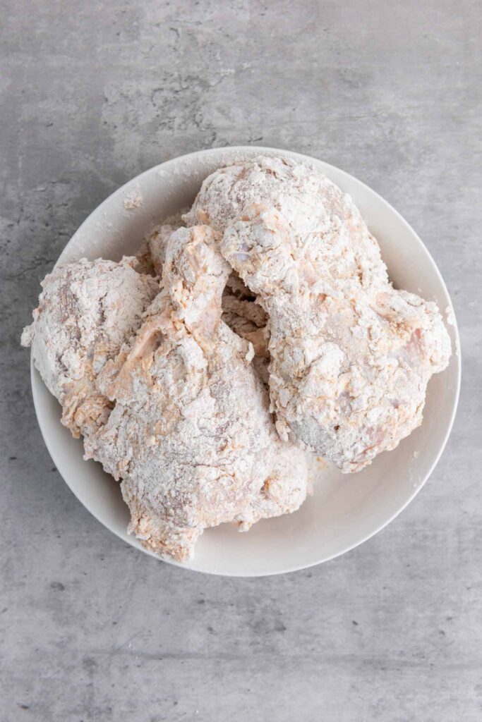 A bowl of buttermilk-marinated, breaded, and floured raw chicken pieces rests on a gray surface.