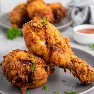 Two pieces of crispy buttermilk fried chicken sit on a gray plate, accompanied by a sauce dish and another plate of delicious chicken in the background.