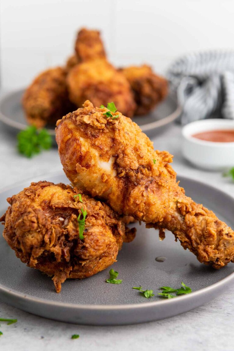 Two pieces of crispy buttermilk fried chicken sit on a gray plate, accompanied by a sauce dish and another plate of delicious chicken in the background.