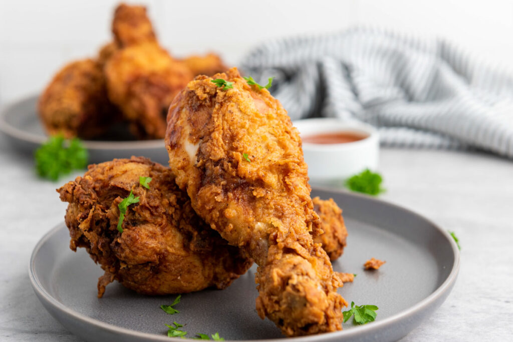 Two pieces of buttermilk fried chicken sit on a gray plate, garnished with parsley, while a striped cloth and dipping sauce add charm to the background.