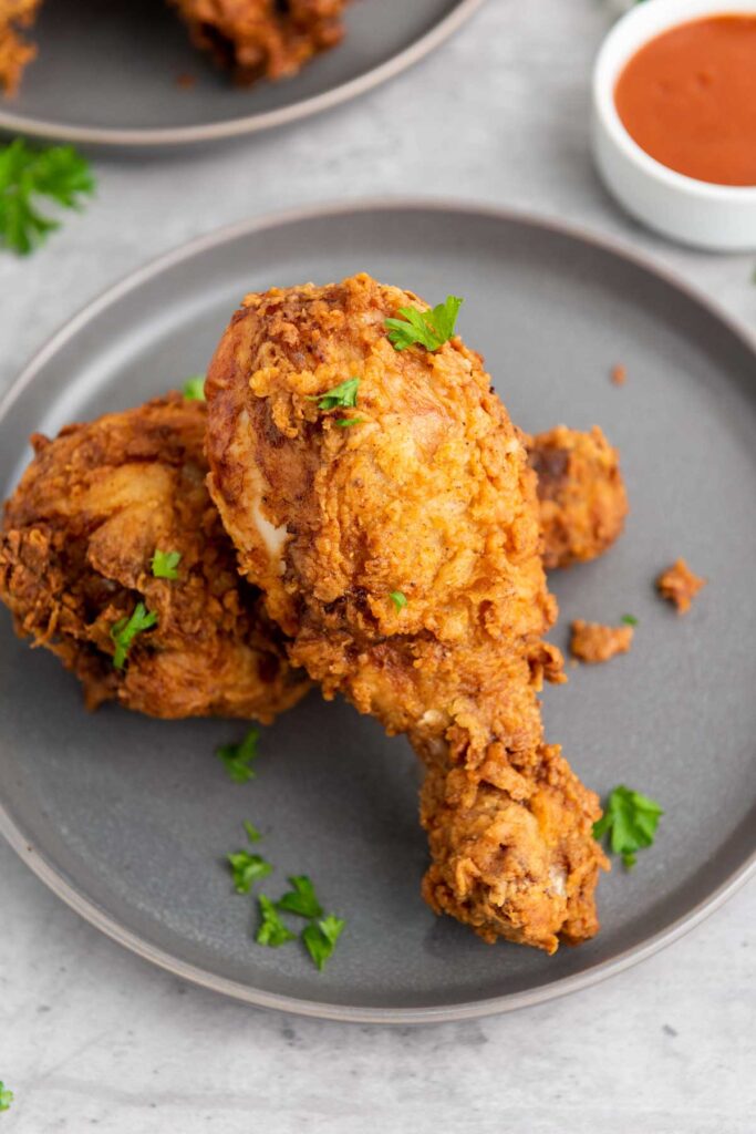 Buttermilk fried chicken drumsticks garnished with parsley rest elegantly on a gray plate, accompanied by a small bowl of dipping sauce in the background.