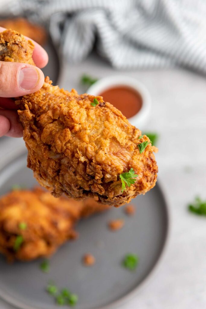 A hand holds a piece of crispy buttermilk fried chicken garnished with parsley, with more chicken and dipping sauce on a gray plate in the background.