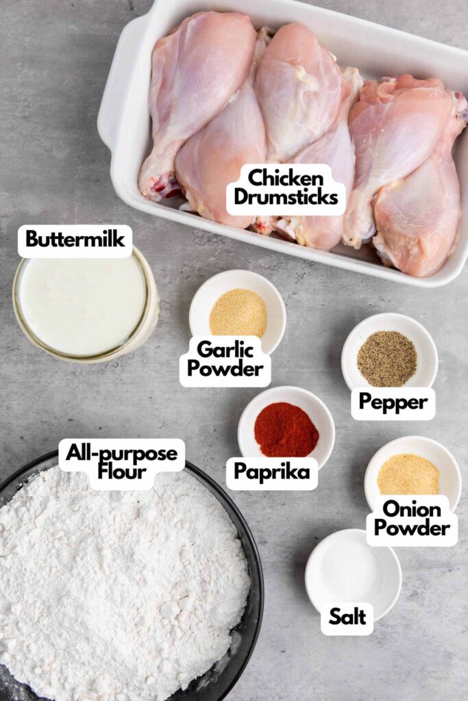 Ingredients for buttermilk fried chicken: raw chicken drumsticks in a dish, a glass of buttermilk, and bowls of all-purpose flour, garlic powder, paprika, pepper, onion powder, and salt.