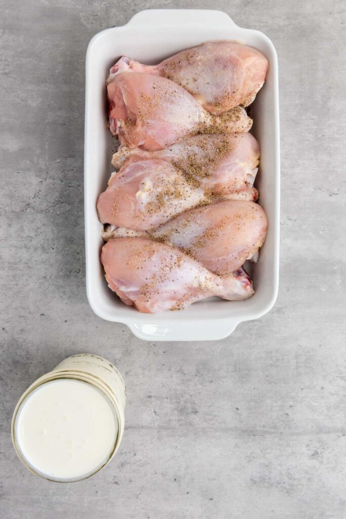 Raw chicken drumsticks in a white baking dish, seasoned with pepper, sit next to a glass of buttermilk on a gray surface, ready to be transformed into crispy fried chicken.