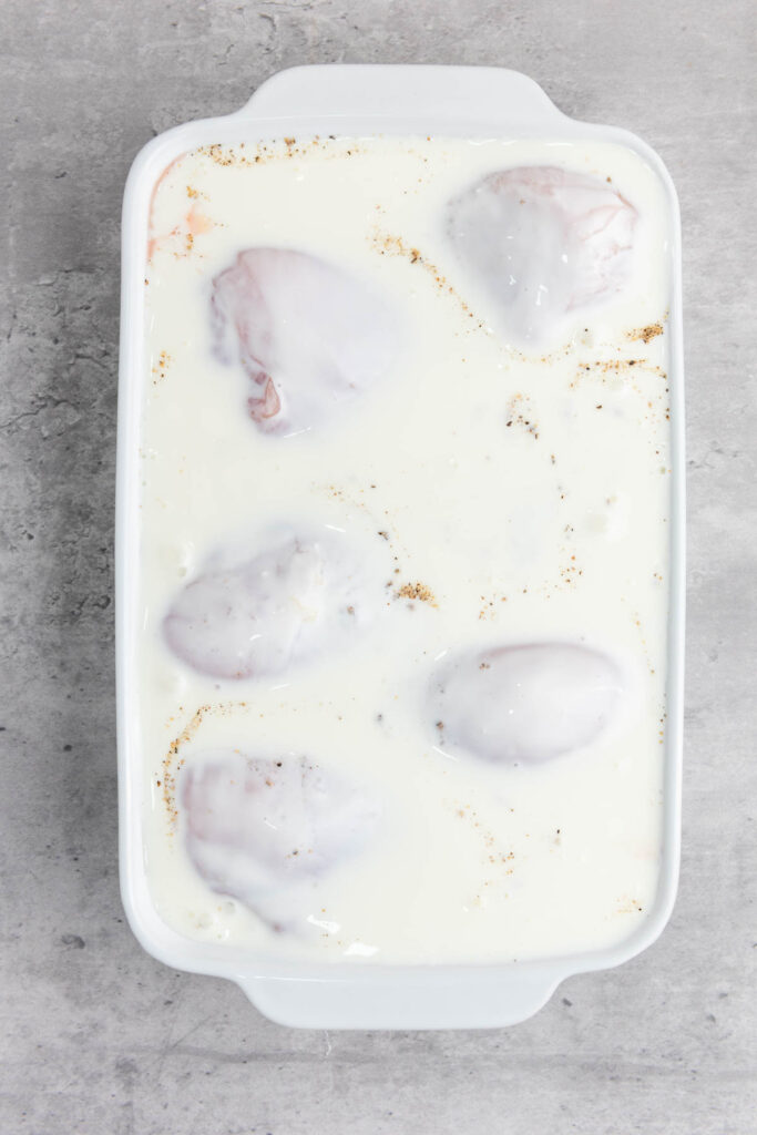 A white baking dish holds five raw chicken pieces marinating in a tangy buttermilk sauce, hinting at the crispy delight of buttermilk fried chicken to come, resting on a gray countertop.