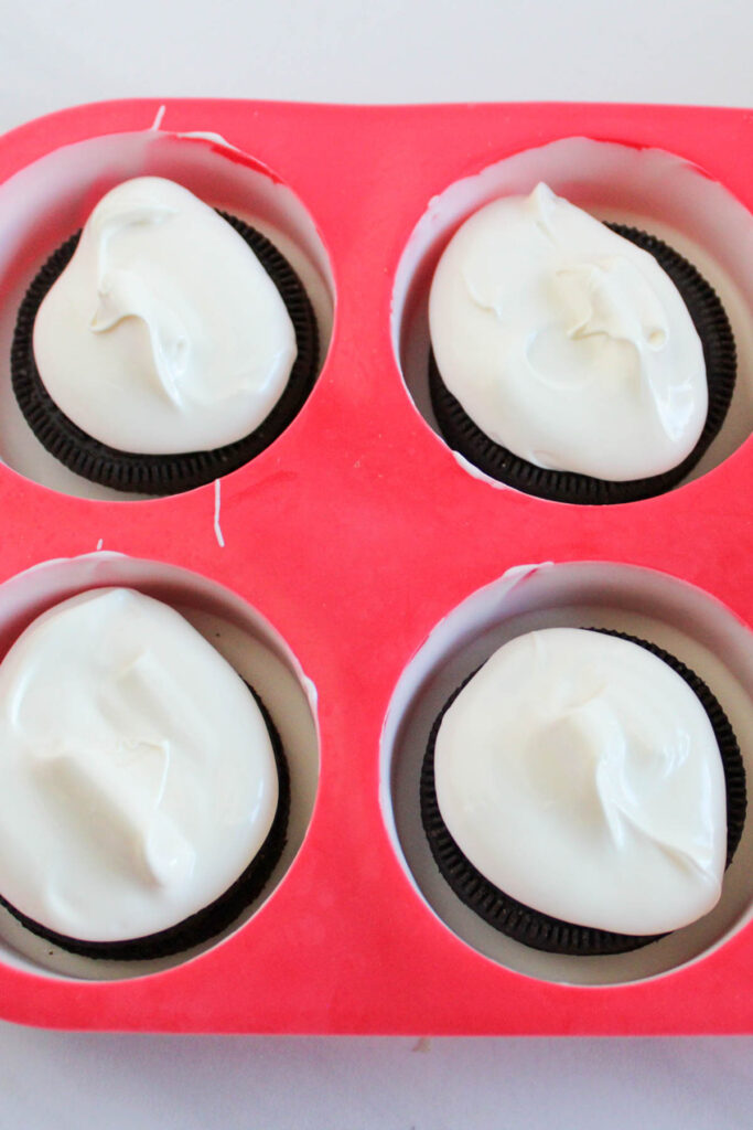 A red silicone mold with four compartments, each containing a chocolate sandwich cookie topped with white cream.