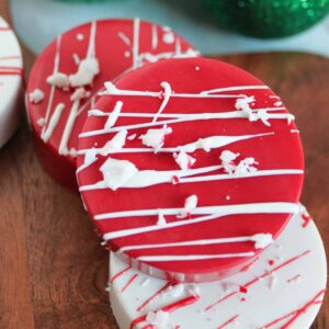Two chocolate-covered cookies, one red and one white, with white chocolate drizzles, placed on a wooden board. Green ornaments are visible in the background.