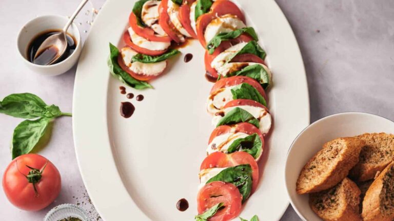 A plate of Caprese salad with sliced tomatoes, mozzarella, and basil leaves, drizzled with balsamic glaze, served with a side of toasted bread.