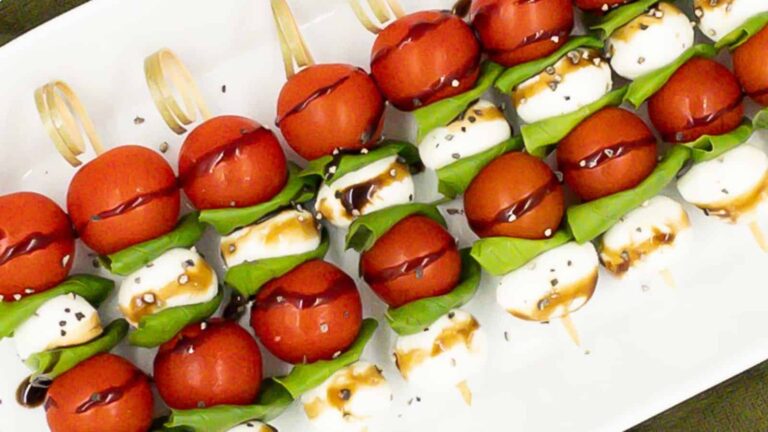 Skewers with cherry tomatoes, mozzarella balls, and basil leaves drizzled with balsamic glaze on a white plate.