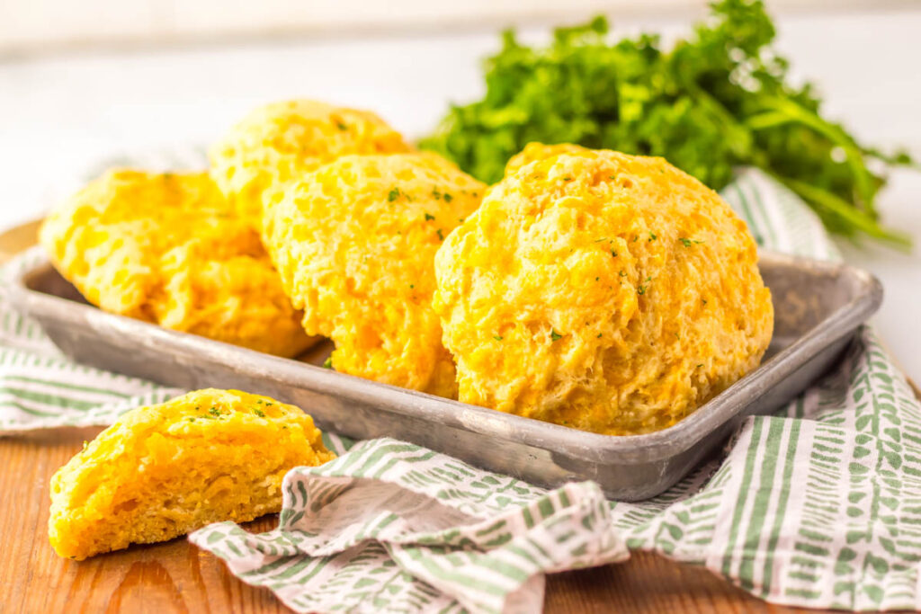 Four golden cheddar bay biscuits on a metal tray with a striped cloth, and a bunch of parsley in the background.