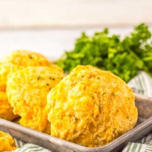 Three golden-brown cheddar bay biscuits rest on a metal tray with a green striped cloth, garnished with parsley in the background.