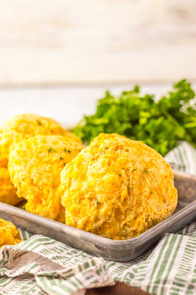 Three golden-brown cheddar bay biscuits rest on a metal tray with a green striped cloth, garnished with parsley in the background.