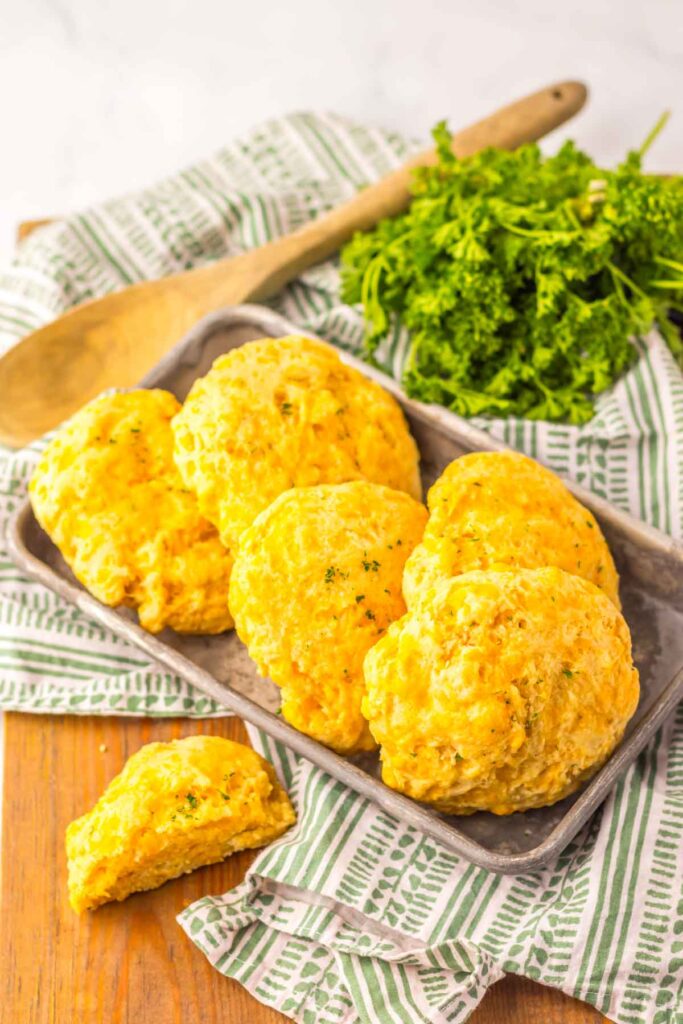 A tray of cheddar bay biscuits sits on a striped cloth, accompanied by a wooden spoon and fresh parsley.