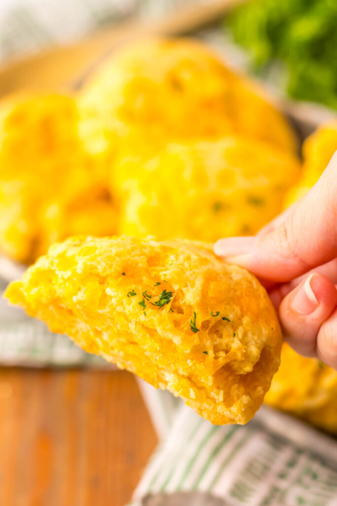 A hand holds a golden cheddar bay biscuit with a bite taken out, revealing the soft, textured inside. Other biscuits are blurred in the background.