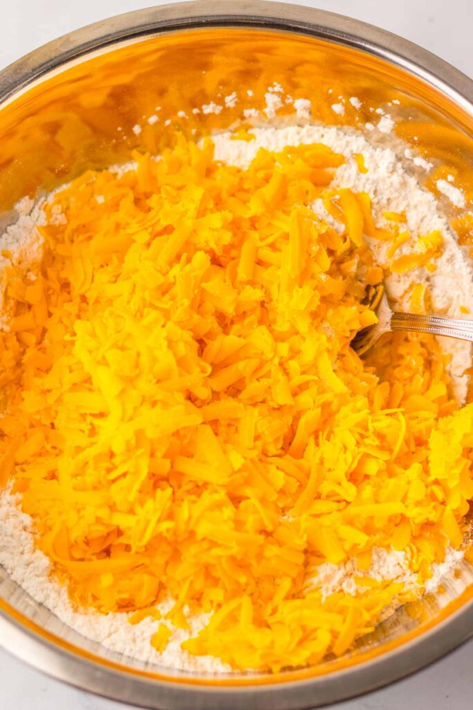 A metal bowl filled with shredded cheddar cheese, hinting at the deliciousness of cheddar bay biscuits in the making, rests atop a bed of flour. A spoon lies partially visible on the side, ready to mix the ingredients into savory perfection.
