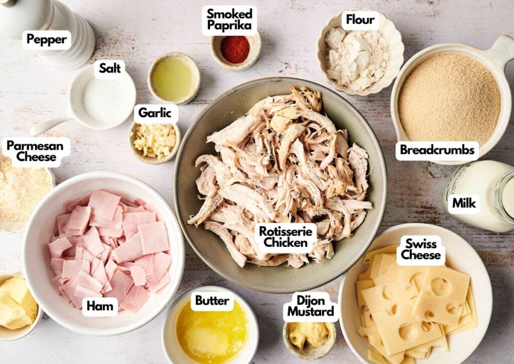 A variety of labeled ingredients on a table, perfect for a Chicken Cordon Bleu Casserole Recipe, including rotisserie chicken, ham, Swiss cheese, breadcrumbs, flour, milk, butter, Dijon mustard, garlic, Parmesan cheese, salt, pepper, and smoked paprika.