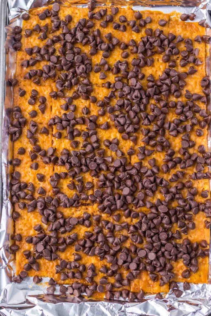A sheet of baked crackers topped with melted chocolate chips and drizzled with strawberry toffee bark on a foil-lined tray.