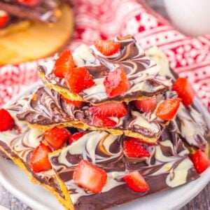 A stack of chocolate covered strawberry toffee bark with a marbled design is elegantly topped with diced strawberries on a white plate. A red patterned cloth adds charm in the background.