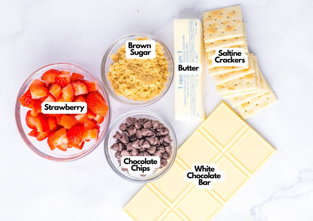 Bowls of chopped strawberries, brown sugar, chocolate chips, and a stick of butter are arranged with saltine crackers and a white chocolate bar on a white surface&mdash;each labeled for making Chocolate Covered Strawberry Toffee Bark.