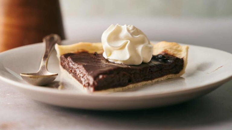 A slice of chocolate pie with whipped cream on top is placed on a plate. A small spoon rests beside the dessert.