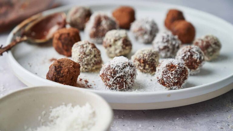 A plate of assorted truffles covered in cocoa and shredded coconut. A spoon and a small bowl with coconut flakes are nearby.