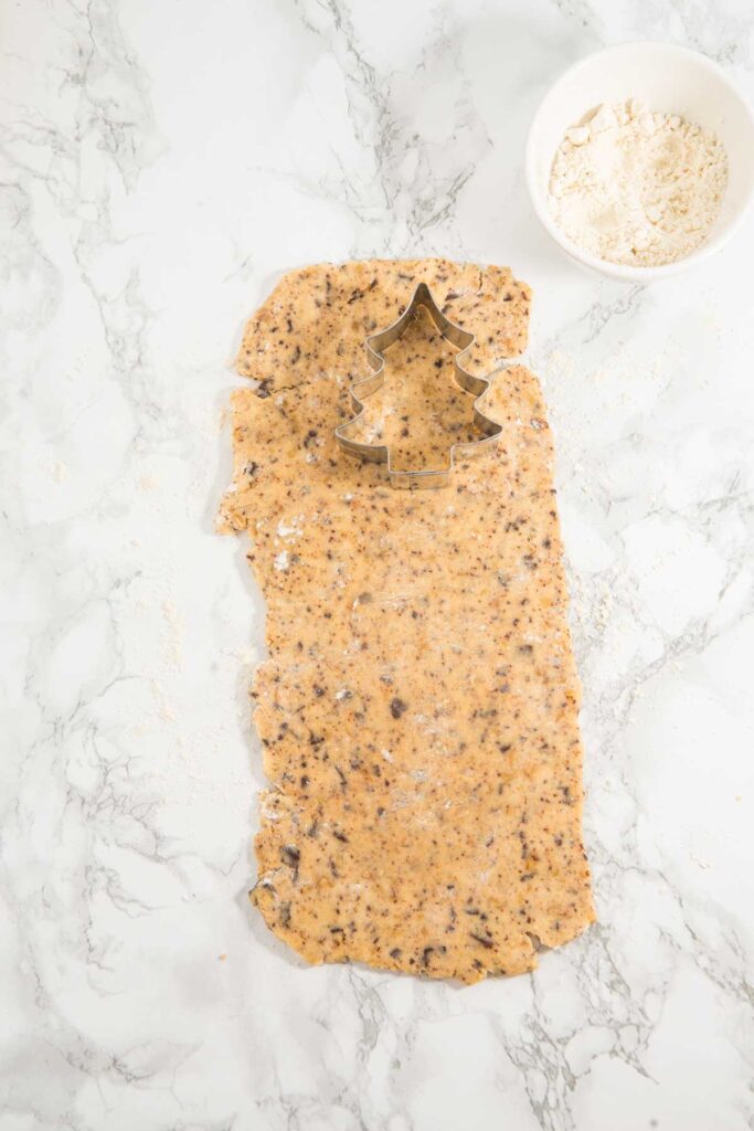 Cookie dough is rolled out on a marble surface with a Christmas tree-shaped cookie cutter, perfect for crafting Christmas tree cookies for hanging. A bowl of flour sits nearby, ready to dust the dough for easy cutting.