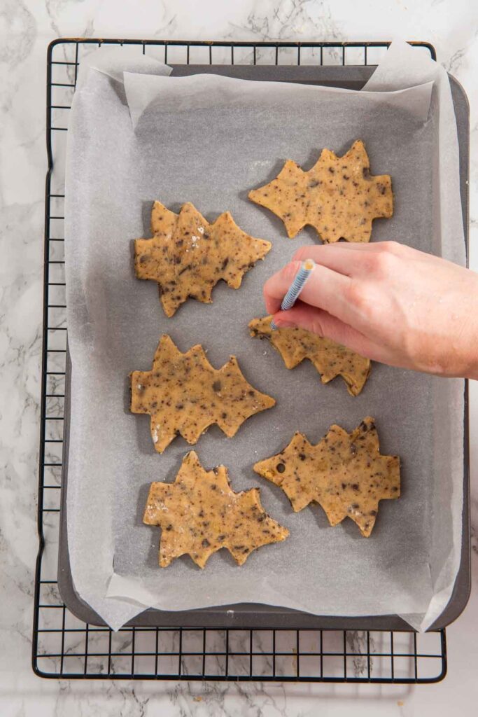A hand carefully pokes holes in leaf-shaped cookie dough pieces on a parchment-lined baking tray, transforming them into delightful Christmas tree cookies for hanging.
