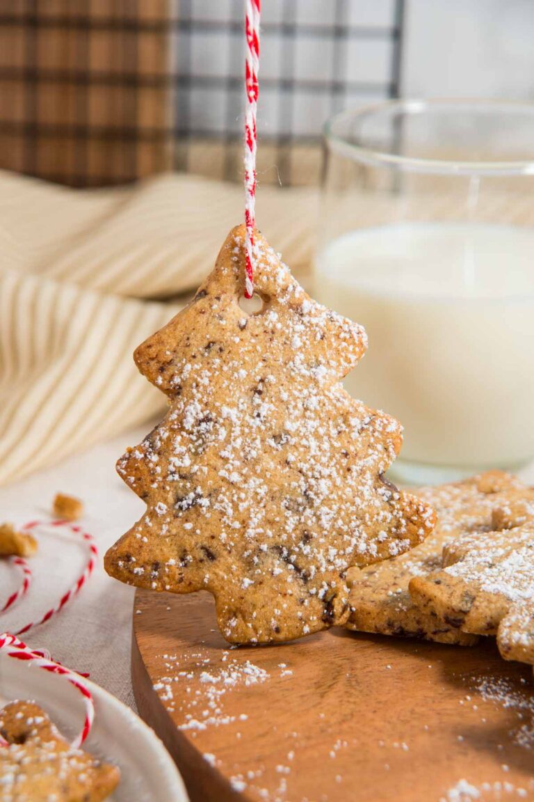 A Christmas tree cookie for hanging, dusted with powdered sugar, dangles by a red-and-white string near a glass of milk on a wooden surface.
