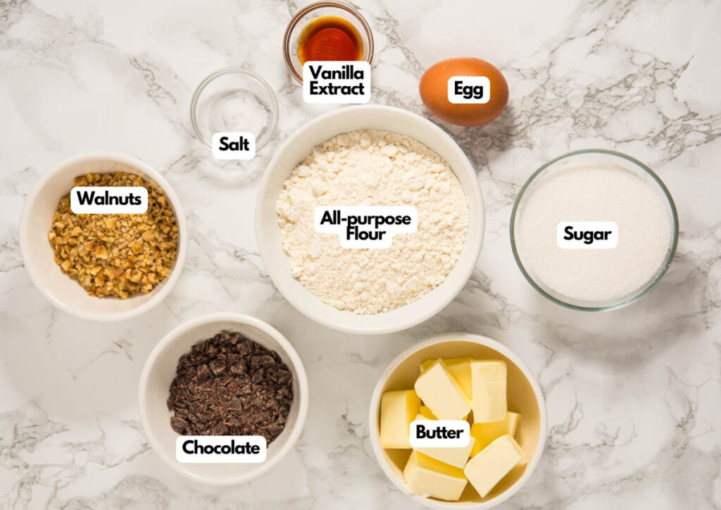 Top-down view of measured baking ingredients in bowls labeled as all-purpose flour, sugar, butter, chocolate, walnuts, salt, vanilla extract, and an egg on a marble surface&mdash;perfect for making Christmas tree cookies for hanging.