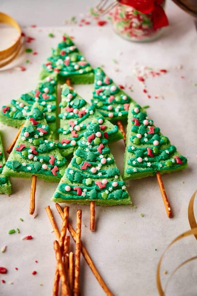 Green Christmas tree-shaped cookies with colorful sprinkles, each on a pretzel stick, arranged on parchment paper.