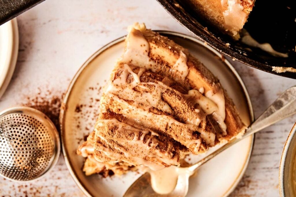 A slice of layered cinnamon roll cake with icing on a plate, sprinkled with cinnamon, sits next to a metal sieve and a spoon.