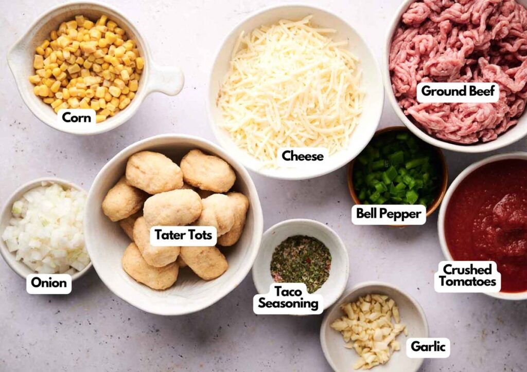 Various labeled bowls containing ingredients for cowboy tater tot casserole&mdash;corn, cheese, ground beef, bell pepper, crushed tomatoes, garlic, taco seasoning, tater tots, and chopped onion&mdash;are arranged on a white surface.