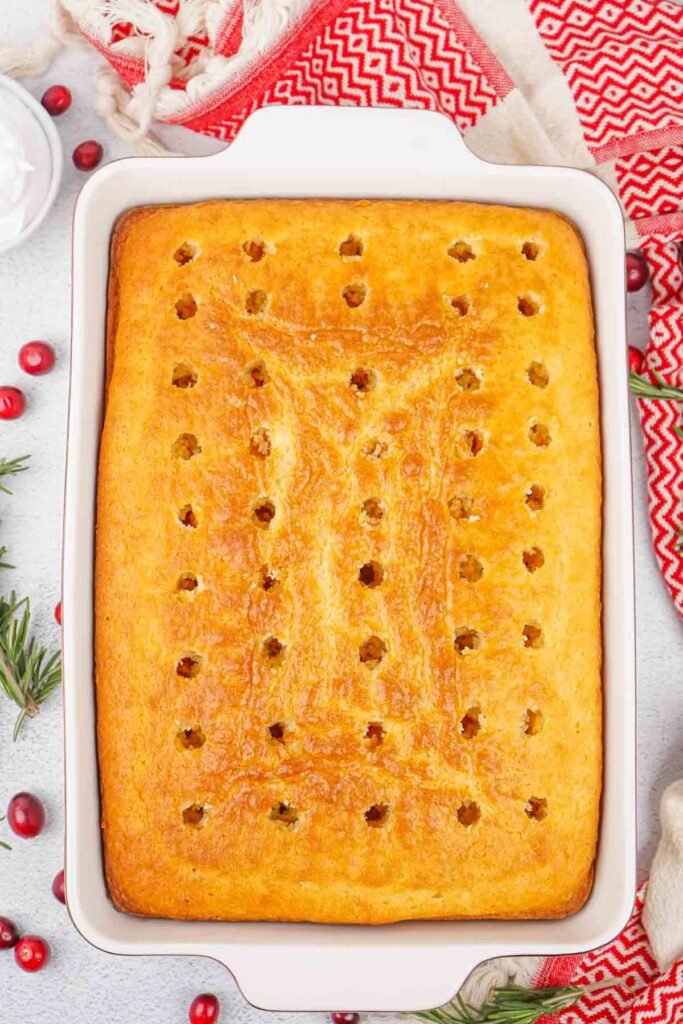 A rectangular Christmas poke cake in a white baking dish, featuring evenly spaced holes on the golden-brown surface, surrounded by cranberries, rosemary, and a red patterned cloth.
