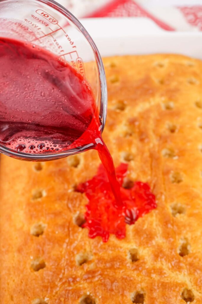 A festive red liquid cascades from a measuring cup onto the Christmas Poke Cake, seeping into its surface holes and infusing it with holiday cheer.