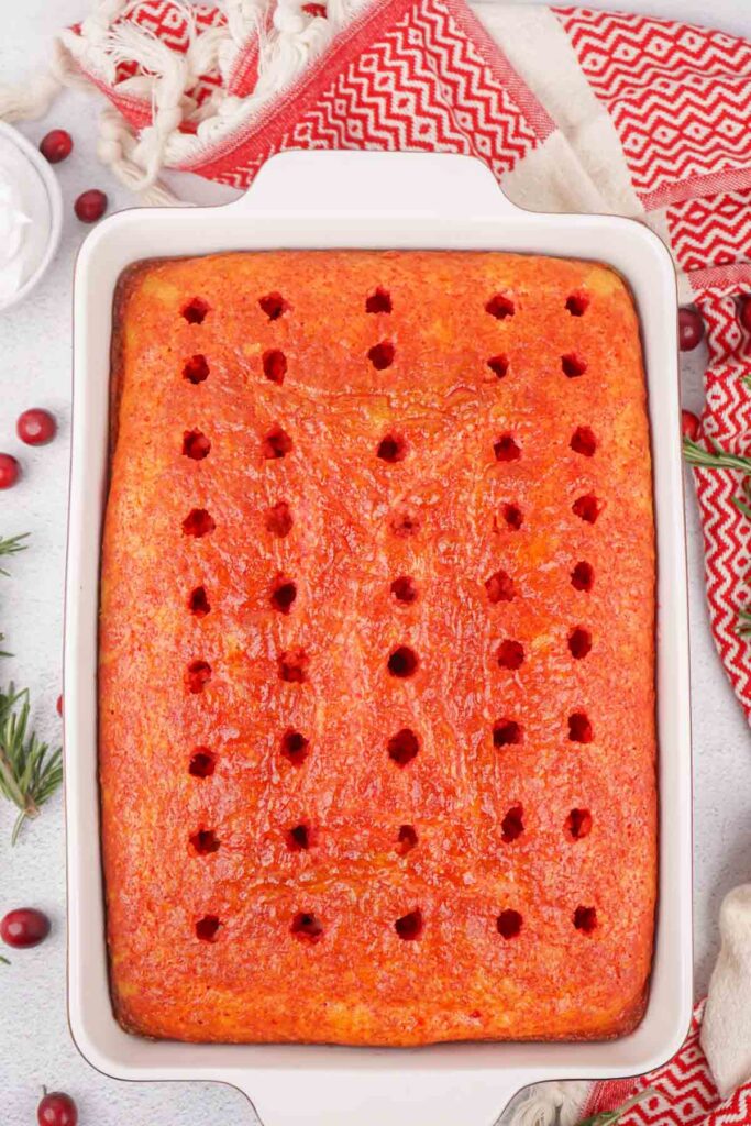 A rectangular Christmas Poke Cake in a baking dish, topped with red glaze and evenly spaced small holes. Nearby are red and white patterned cloths, cranberries, and rosemary sprigs.