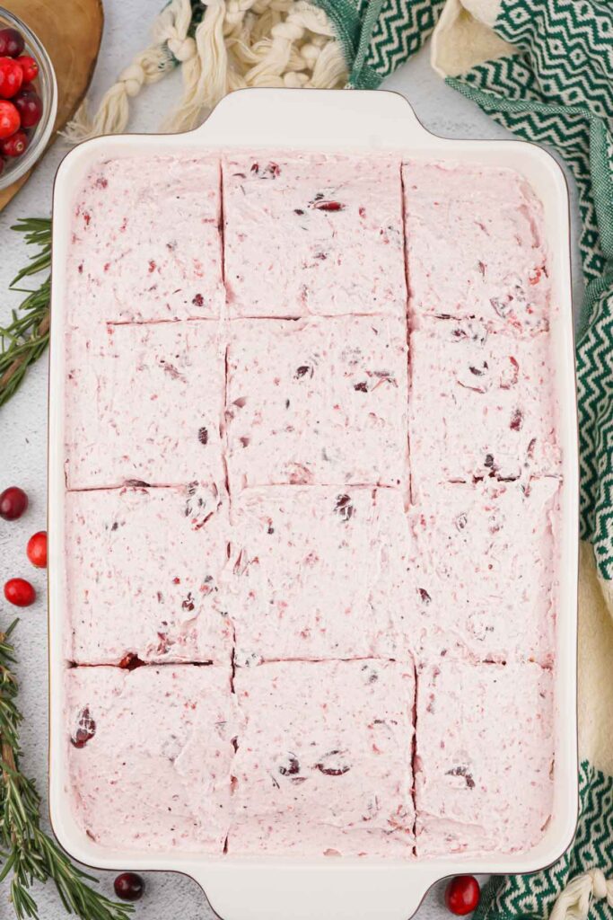 A festive dish of sliced cranberry salad with creamy topping, beautifully surrounded by rosemary sprigs and cranberries, reminiscent of a Christmas poke cake.