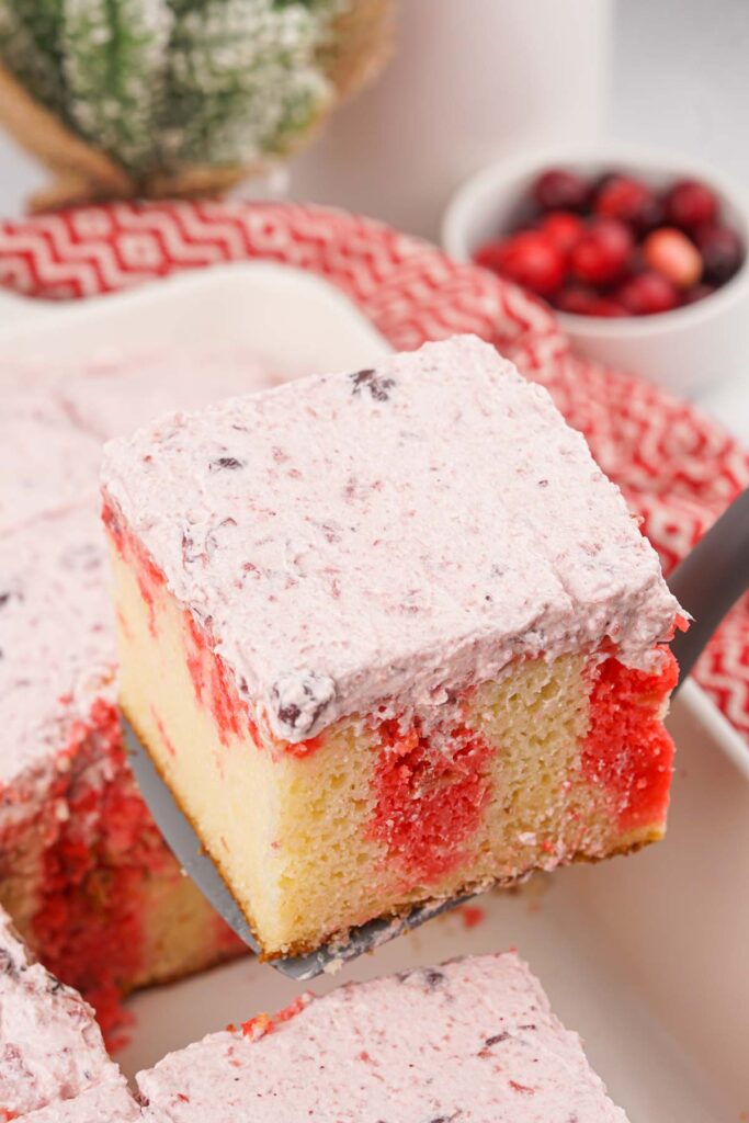 A slice of Christmas Poke Cake with vanilla layers, red streaks, and pink frosting is being lifted from a baking dish. In the background, a bowl of cranberries and a festive red-patterned napkin enhance the holiday spirit.