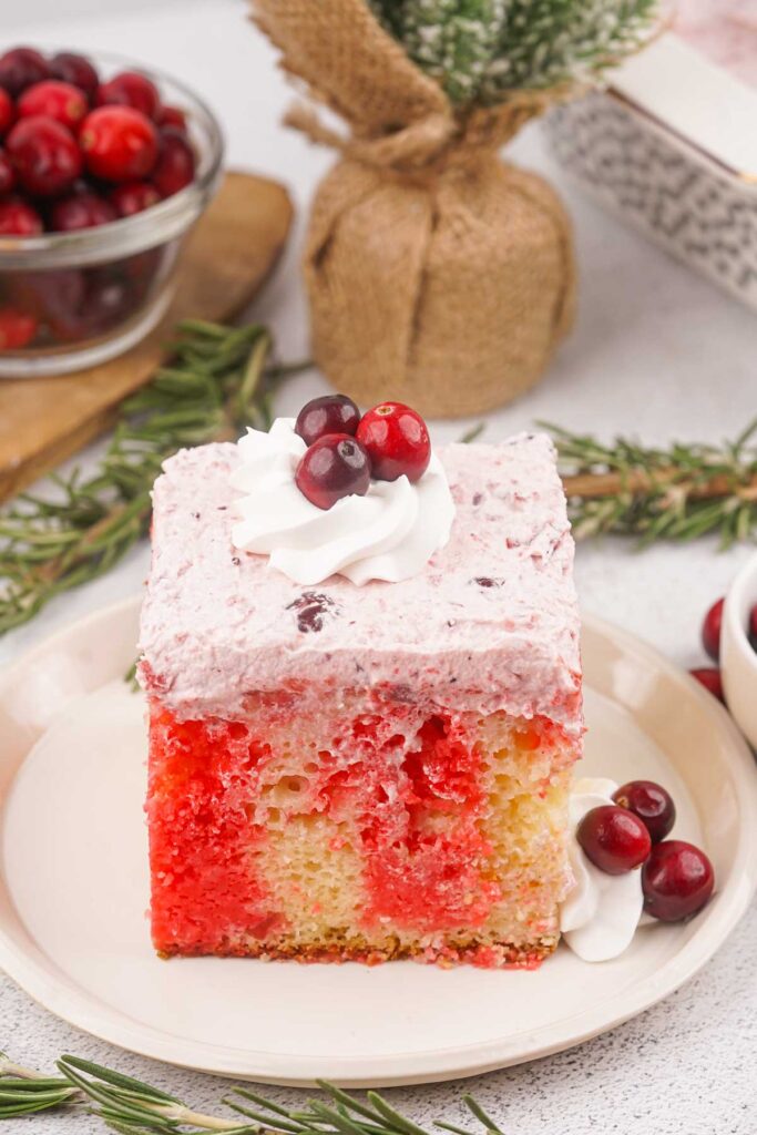 Close-up of a Christmas Poke Cake featuring layers of red and yellow cake topped with whipped cream and cranberries, all beautifully presented on a plate. Fresh cranberries sit beside it, enhanced by a small plant and sprigs of rosemary for decoration.