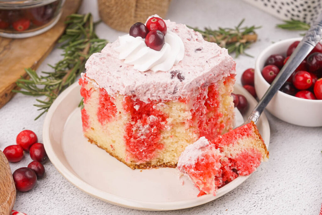 A slice of Christmas Poke Cake with red swirls sits elegantly, topped with pink frosting, whipped cream, and cranberries on a plate with a fork. Nearby are sprigs of rosemary and a bowl of fresh cranberries, enhancing the festive charm.