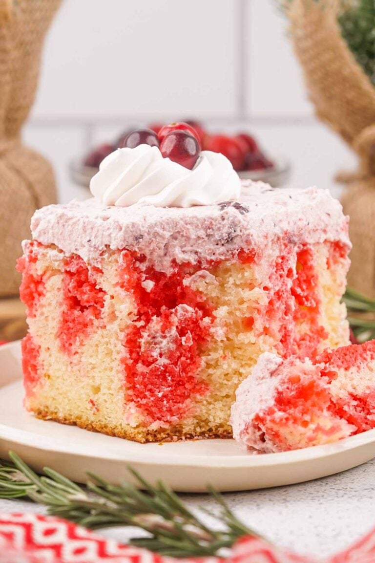 A slice of Christmas Poke Cake with vibrant red and yellow layers, topped with whipped cream and a cherry, rests on a white plate.