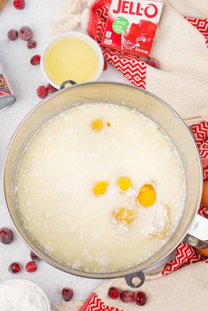 A large bowl containing a mixture of eggs, flour, and liquid ingredients sits on a kitchen counter, ready to become a festive Christmas Poke Cake. It's surrounded by cranberries, a Jell-O box, and a red-patterned cloth.