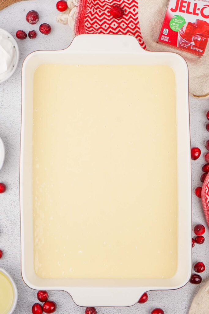 A rectangular baking dish with smooth yellow batter, perfect for a festive Christmas Poke Cake, surrounded by cranberries, a Jell-O box, cream bowls, and a red textured cloth.