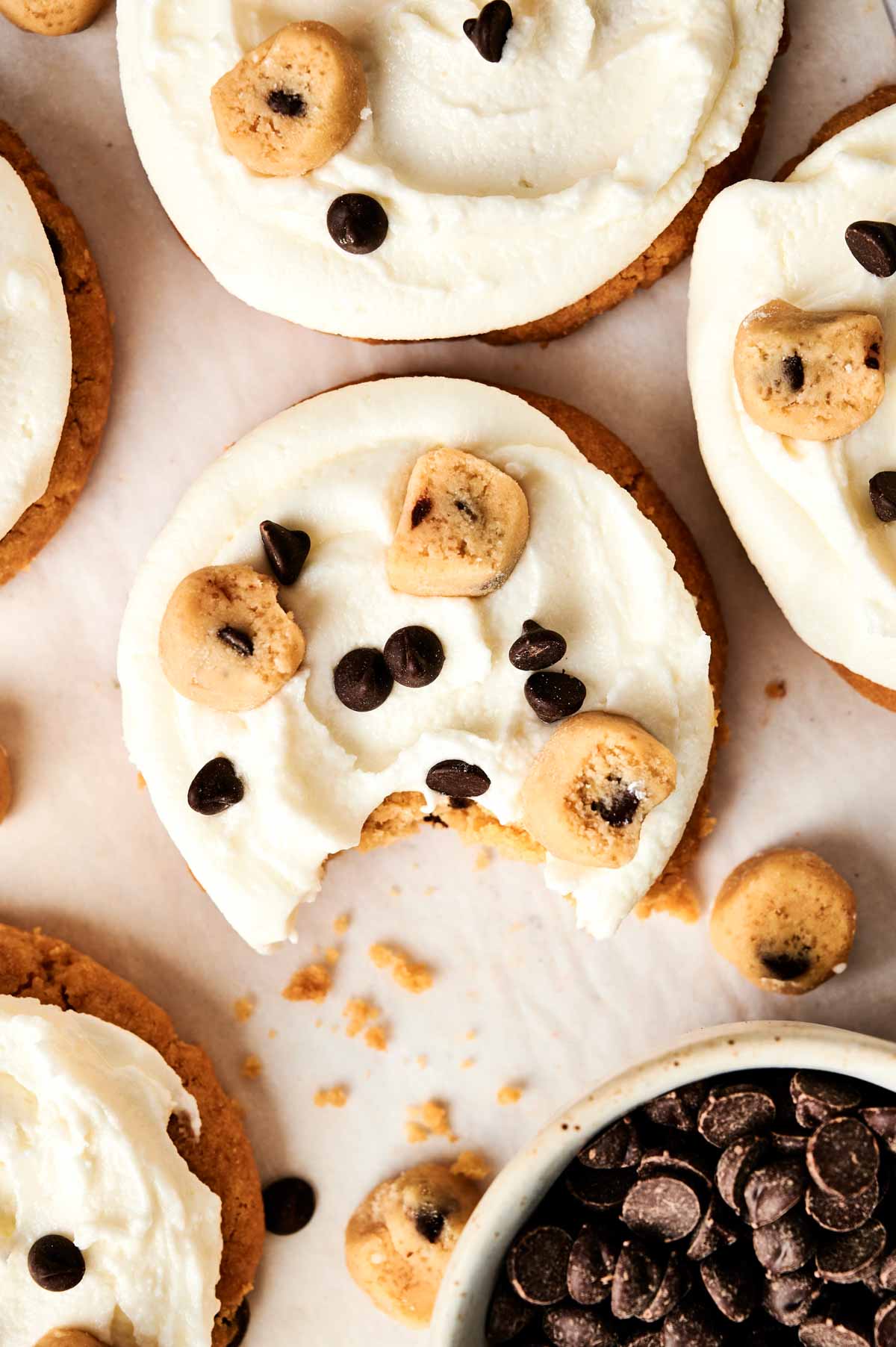 A cookie with white frosting, topped with chocolate chips and cookie dough bites, has a bite taken out. Other similar cookies surround it, with a bowl of chocolate chips nearby.