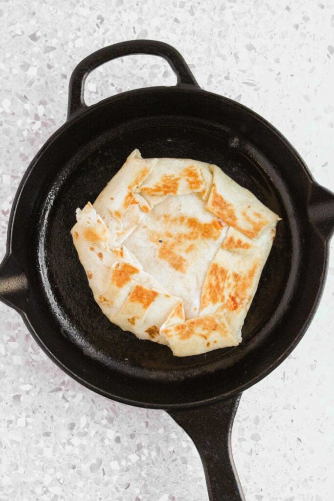 A folded, grilled tortilla wrap is cooking in a black cast iron skillet placed on a speckled countertop.