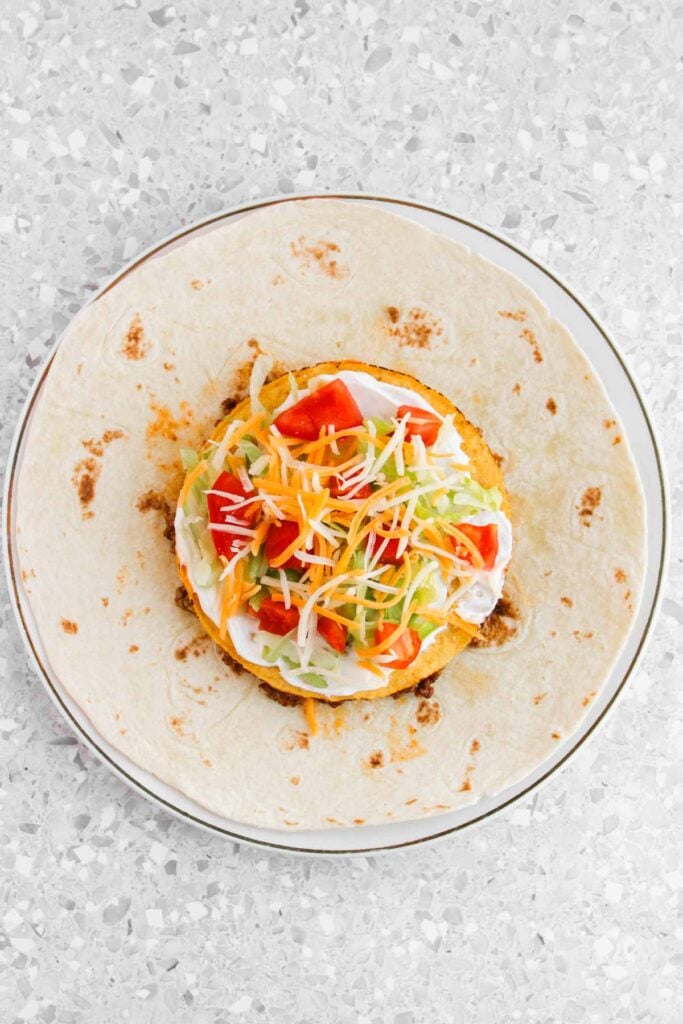 A tortilla with a crispy patty topped with shredded lettuce, tomatoes, carrots, and grated cheese on a plate.